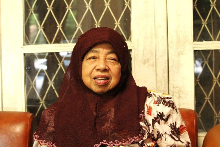 Older woman wearing a maroon hijab and patterned blouse sits on a brown leather couch by a window with lattice panes.