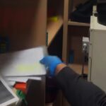 Worker wearing blue gloves reaching into a cabinet beside a metal machine in a dim workplace scene