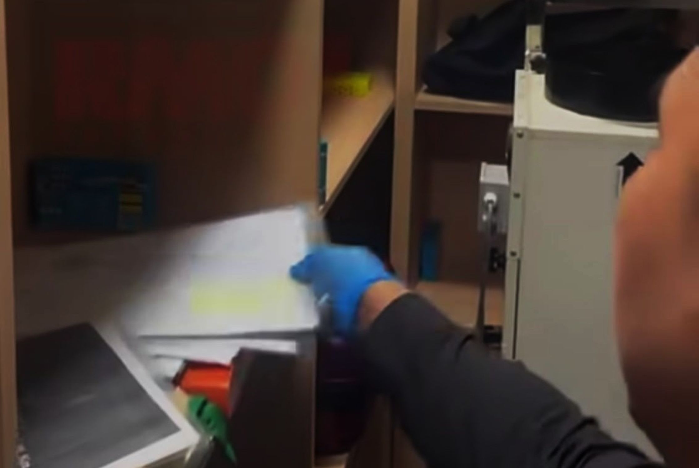 8gdokumen catrawarta Worker wearing blue gloves reaching into a cabinet beside a metal machine in a dim workplace scene