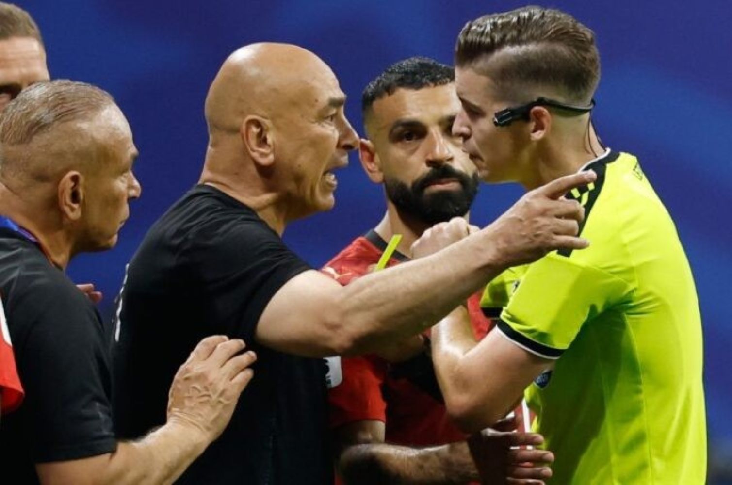 8gmesir1 catrawarta Tense moment on a soccer field as a bald man in black shouts at a goalkeeper in a bright yellow jersey with teammates watching closely