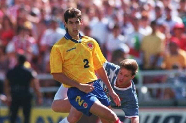 Soccer player in a yellow jersey (#2) sprinting with the ball while a blue-jersey opponent challenges him.