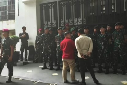 Group of soldiers in uniform forming a line outside a gated building, while two men face them in front.