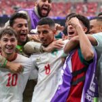 A group of soccer players in white jerseys celebrate together on the field arms around each other in joy after a victory