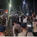 Nighttime street scene with a crowd gathered on the sidewalk including several uniformed officers or security personnel and cars parked along the curb