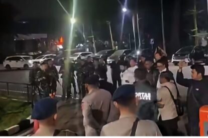 Nighttime street scene with a crowd gathered on the sidewalk, including several uniformed officers or security personnel, and cars parked along the curb.