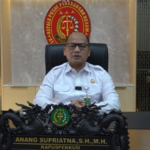 Man in a white uniform shirt sits at a dark carved desk in a formal office with a government emblem behind him