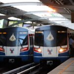 Two bts skytrain cars are parked at an elevated station platform with passengers boarding nearby and metal overhead beams above