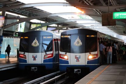 Two bts skytrain cars are parked at an elevated station platform with passengers boarding nearby and metal overhead beams above