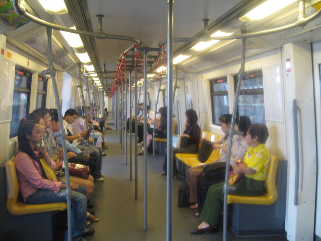 Interior of a subway car with passengers seated along both sides and red overhead hand straps overhead