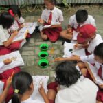 Group of schoolchildren in uniforms sitting on the ground in a circle painting on paper with orange brushes and bowls of black paint nearby