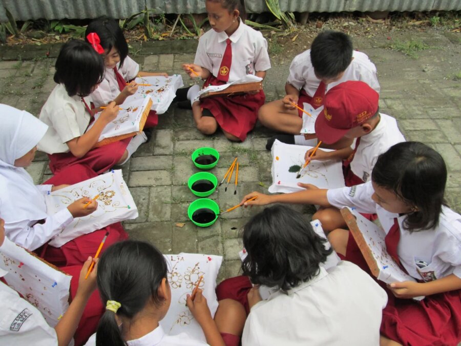 Belajarsambilmenggambare1783143361842 catrawarta Group of schoolchildren in uniforms sitting on the ground in a circle painting on paper with orange brushes and bowls of black paint nearby
