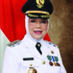 Portrait of a woman in a white ceremonial uniform with medals and badges wearing a hijab and peaked cap in front of the indonesian flag