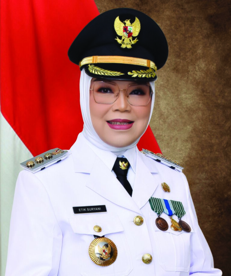 Portrait of a woman in a white ceremonial uniform with medals and badges, wearing a hijab and peaked cap, in front of the Indonesian flag.
