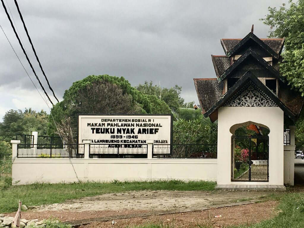 Front view of a white wall and ornate gate with a large sign reading'Departemen Sosial RI, Makam Pahlawan Nasional Teuku Nyak Arief, 1899-1946' against a cloudy sky.