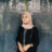 Smiling woman wearing a beige hijab, black jacket, striped shirt, and a watch, standing against a dark textured wall.