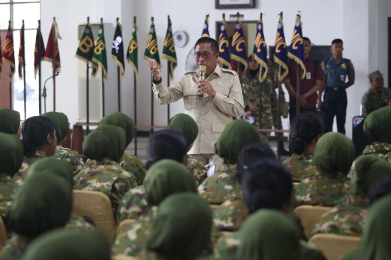 A man in a tan military uniform speaks into a microphone to a roomful of soldiers in camouflage, with flags lined up in the background.