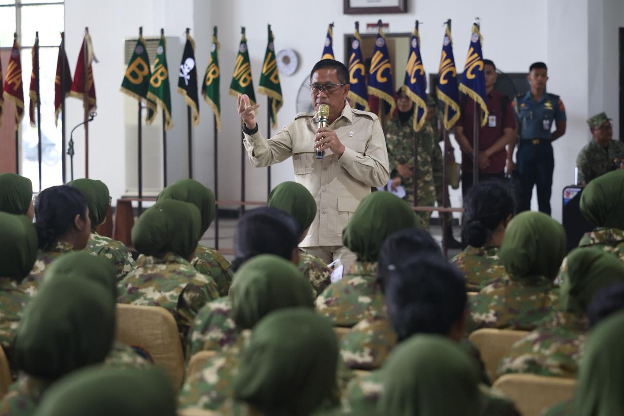 Donny ermawan catrawarta A man in a tan military uniform speaks into a microphone to a roomful of soldiers in camouflage with flags lined up in the background