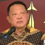 Man in a patterned batik shirt speaks into a microphone at a podium during a formal event with a red and gold backdrop behind him