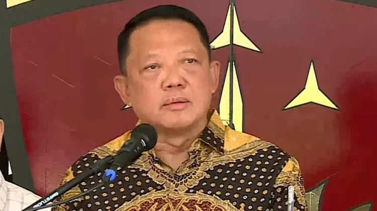 Febrie catrawarta Man in a patterned batik shirt speaks into a microphone at a podium during a formal event with a red and gold backdrop behind him