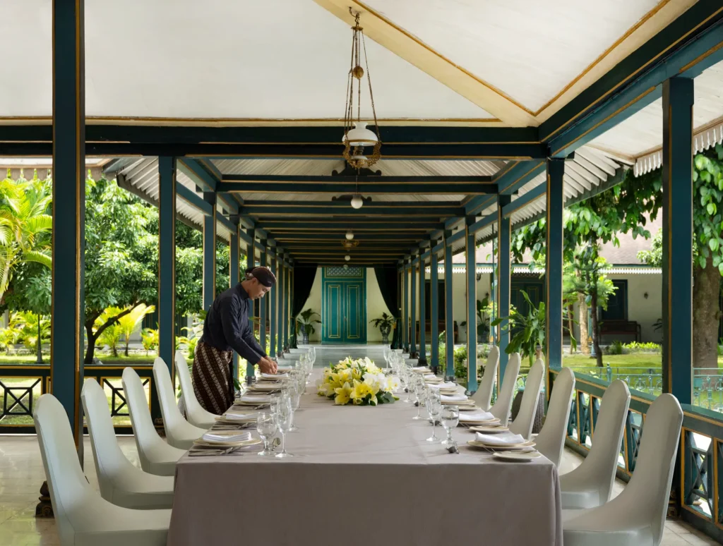 Gadrilunch catrawarta Long banquet table set for a formal dinner under a teal framed covered walkway white chairs glassware and a yellow floral centerpiece
