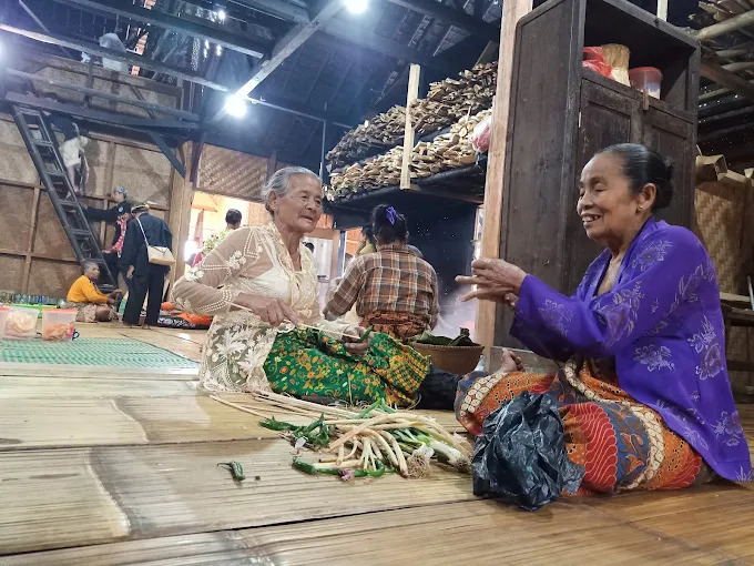 Two elderly women sit on a wooden floor in a rustic market chatting while arranging fresh lemongrass stalks for sale