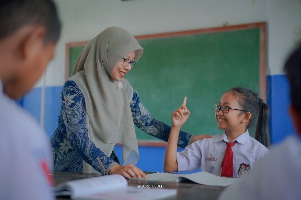 Haribelajargurukebijakankemendikdasmendenganjadwalbelajarseminggu catrawarta Teacher in hijab helping a smiling student who raises her finger in a classroom with a green chalkboard behind them