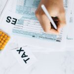 Person writes on tax forms at a marble desk with a calculator nearby and a tax card visible