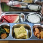 Person holding a metal bento style lunch tray with watermelon rice greens with carrot fried tofu and meat stew in separate compartments
