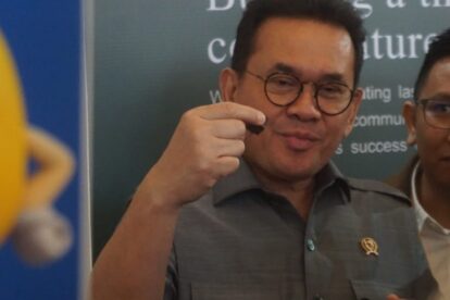 Man in gray shirt and glasses holds up a small dark object between his fingers during a event with a badge visible on his chest and a backdrop in the background