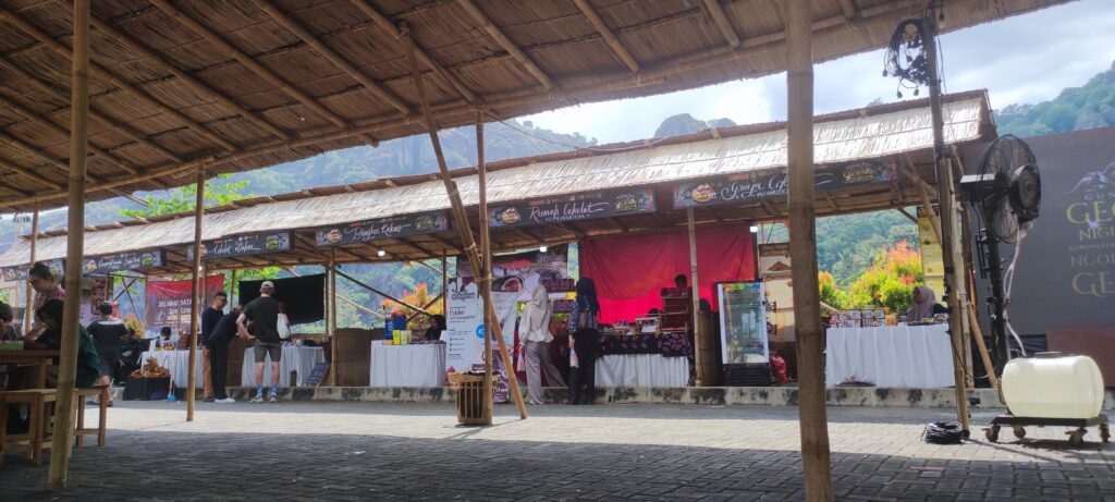 Img20260711115740 catrawarta Open air market with row of wooden stalls under a thatched roof people browsing and chatting against a mountain backdrop