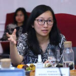Woman with glasses speaking and gesturing at a conference table others seated in red chairs in the background