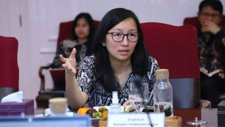 Woman with glasses speaking and gesturing at a conference table, others seated in red chairs in the background.