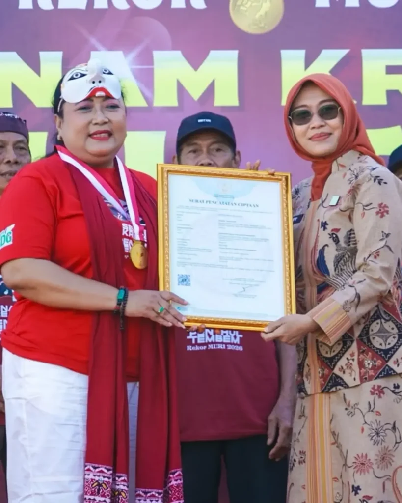 Two women on a stage exchange a framed certificate during an award ceremony one in red with a decorative mask and medal the other in a beige patterned dress with an orange hijab against a purple backdrop
