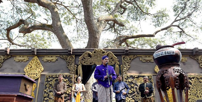 Outdoor stage performance with a singer in a blue jacket and traditional attire flanked by ensemble performers ornate gold backdrop under a large tree a decorative drum on the right