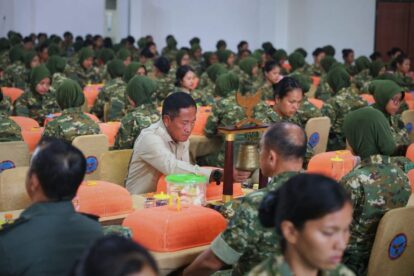 A man in a light uniform rings a ceremonial bell while surrounded by rows of soldiers in camouflage seated at orange-covered desks in a large hall.