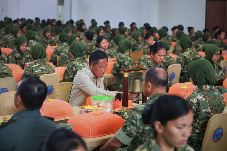 Kemhan catrawarta A man in a light uniform rings a ceremonial bell while surrounded by rows of soldiers in camouflage seated at orange covered desks in a large hall