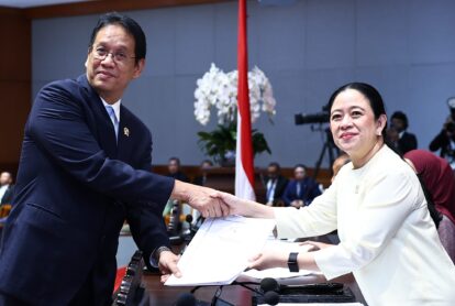 Two officials shake hands across a desk with documents in a formal meeting room, a flag and flowers in the background.