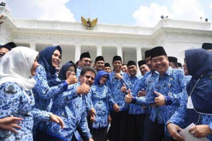 Group of Indonesian officials in blue batik shirts giving thumbs up in front of a white government building with the Garuda emblem.