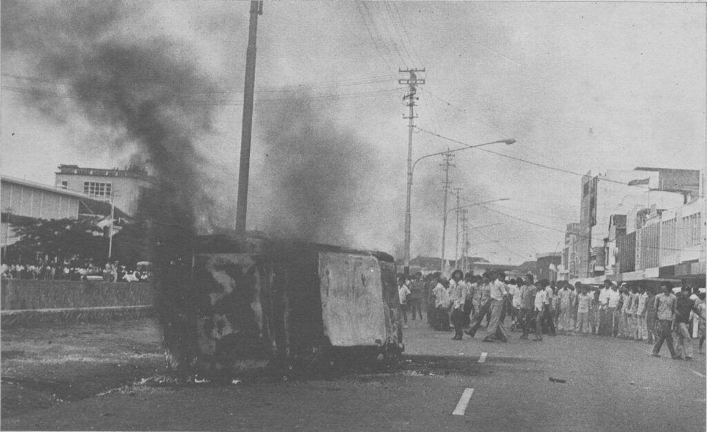 A line of uniformed marchers marching down a city street as a large plume of smoke rises from a building behind them