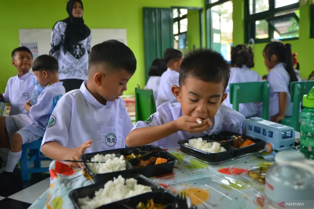 Mbgantara catrawarta Young students in white uniforms eat lunch at a colorful table in a bright green classroom with a teacher standing in the background