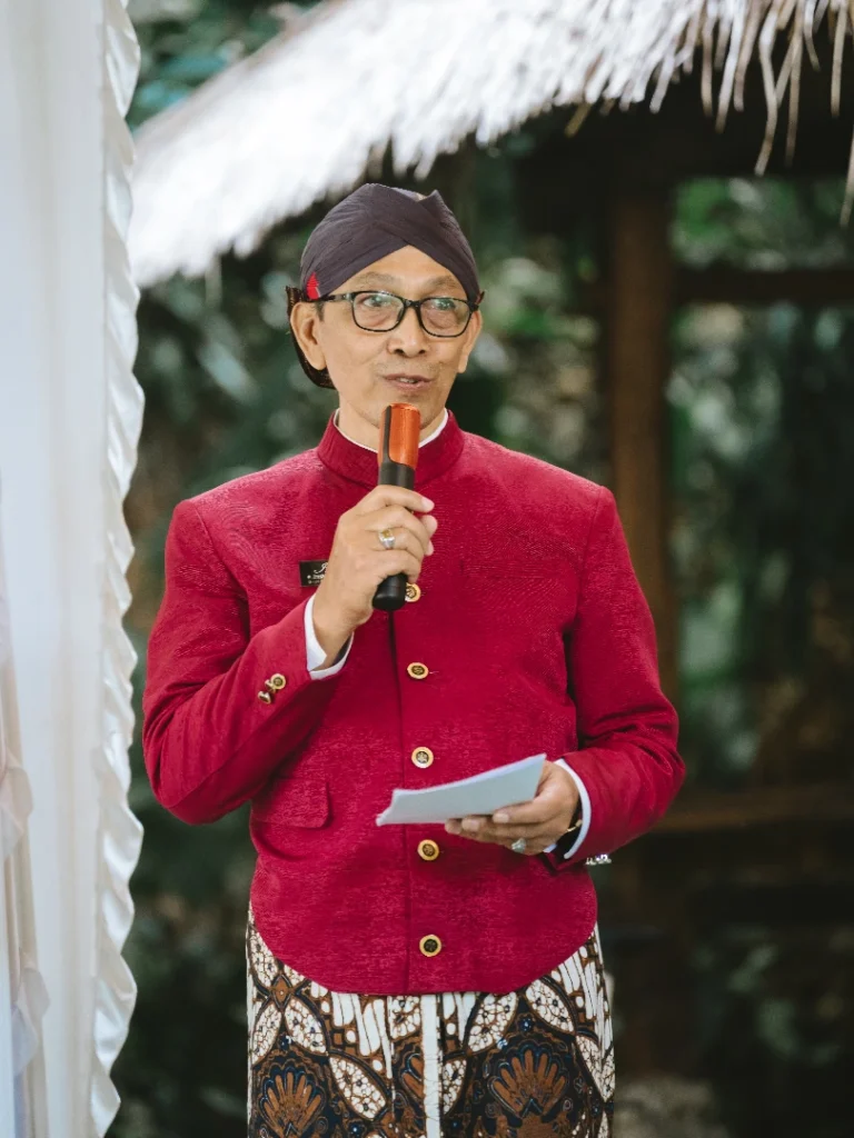 Man in a red traditional jacket and patterned sarong speaks into a handheld microphone outdoors holding notes