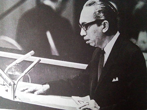 Man in a suit and glasses speaks at a podium reading papers into a microphone