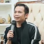 Man in a black jacket with white stripes holding a microphone speaking during an interview while seated on a tufted beige chair in a cozy setting
