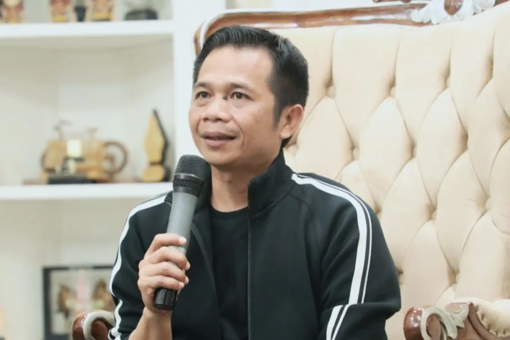 Omzein catrawarta Man in a black jacket with white stripes holding a microphone speaking during an interview while seated on a tufted beige chair in a cozy setting