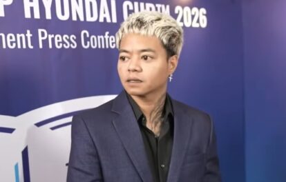 Person in a dark blue suit and black shirt stands against a blue backdrop reading hyundai and press conference 2026 at a formal event they look to the side with an earring visible in one ear