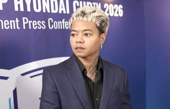 Person in a dark blue suit and black shirt stands against a blue backdrop reading 'HYUNDAI' and 'Press Conference 2026' at a formal event; they look to the side with an earring visible in one ear.