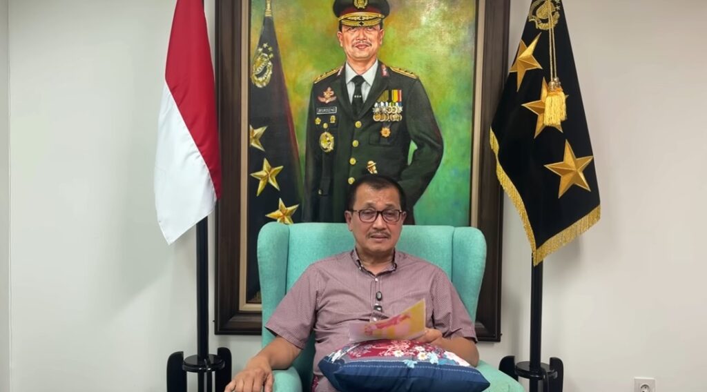 Man in a pink shirt with glasses seated in a teal chair reading papers flanked by indonesian flag and a black flag with gold stars portrait of a military officer behind informative