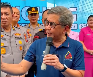 Screenshot 3764 catrawarta Man with glasses in a blue polo speaks into a handheld microphone at a police event with officers and officials in the background