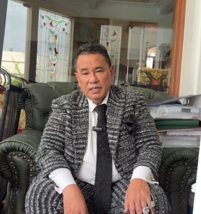 Man in a gray patterned suit and tie sits on a green leather sofa in a room with glass doors and stacks of papers.