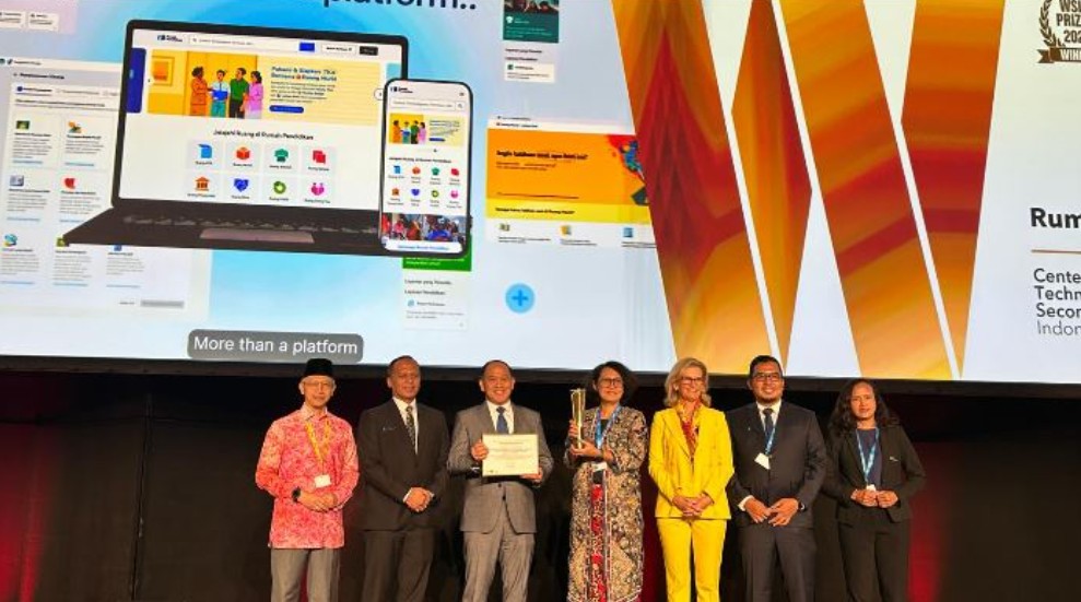 Seven people in business attire on a stage pose with a trophy a large screen displaying devices in the background behind them